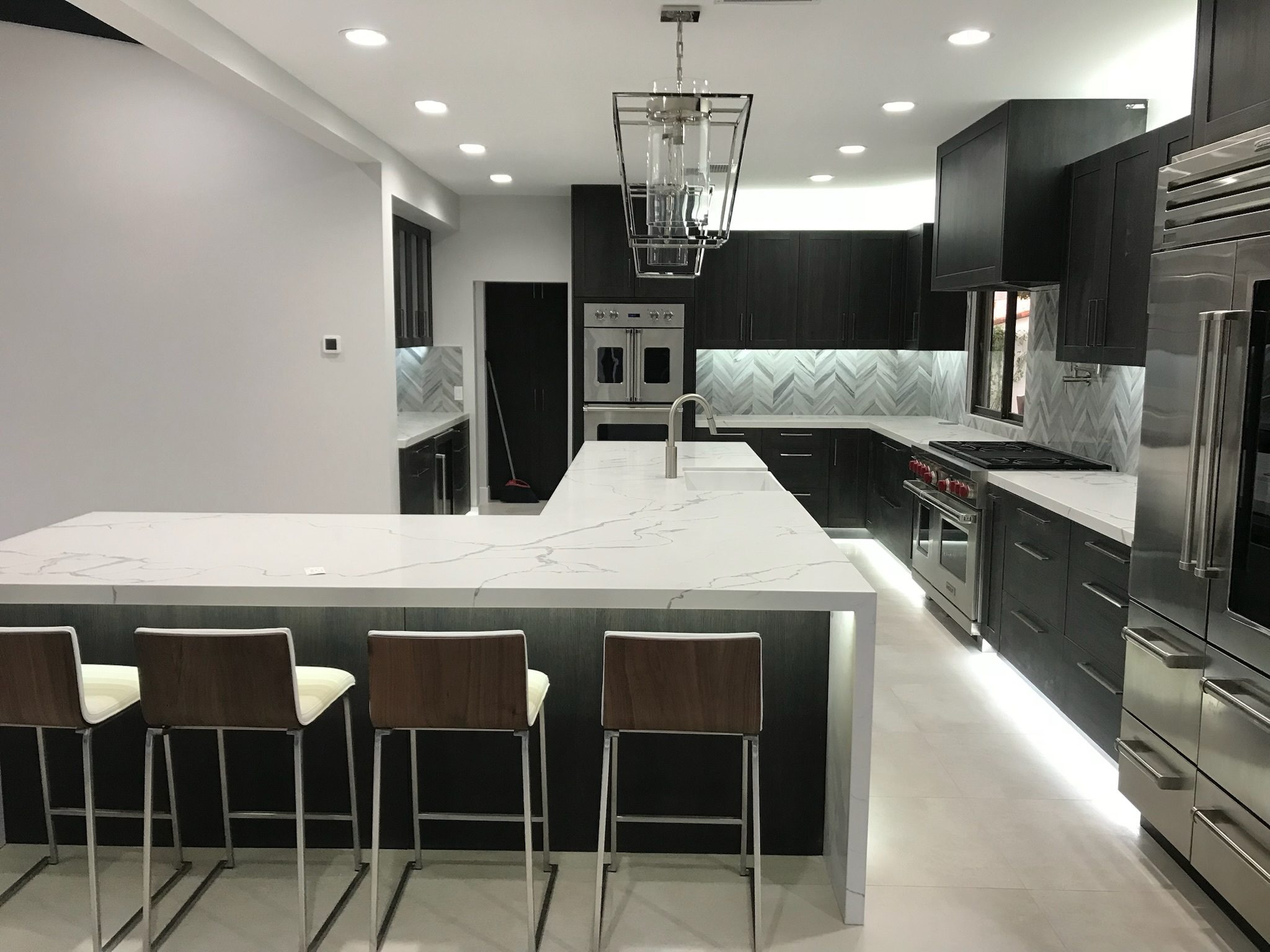 Modern kitchen interior featuring dark wood cabinets, white marble countertops, stainless steel appliances, and a central island with bar stools.